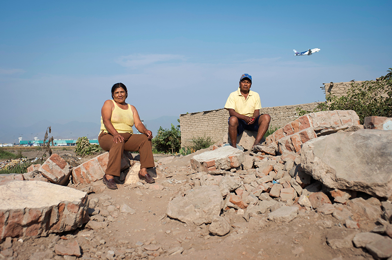 Catalina Guzman Harrimache and her husband Teofilo Huaman Loayza sit on the remnants of a home. The home had its own ground water supply that the town now frequents to wash clothes while sitting amidst the rubble. In the background the main terminal of Jorge Chavez International Airport can be seen. (Photo by Oscar Durand)