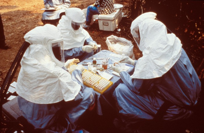 This 1995 photograph shows scientist with personal protective equipment (PPE) testing samples from animals collected in Zaire for the Ebola virus. (Public domain photo via Wikipedia)